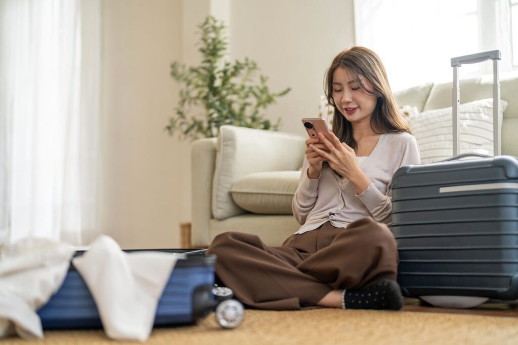 Junge Frau sitzt mit Koffer im Wohnzimmer und bucht ihren Flughafen Shuttle Frankfurt über das Handy